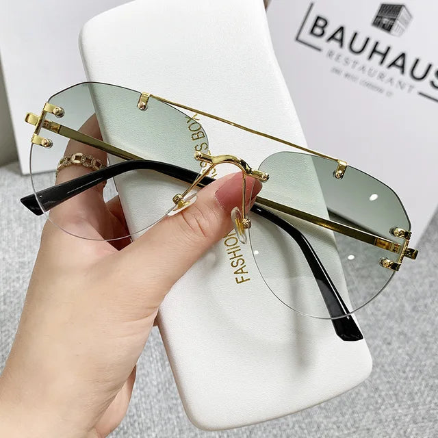 A hand holds stylish eprolo Vintage Rimless Alloy Gradient Pilot Sunglasses, perfect for eye protection, above a white case. In the background, BAUHAUS RESTAURANT is blurred.