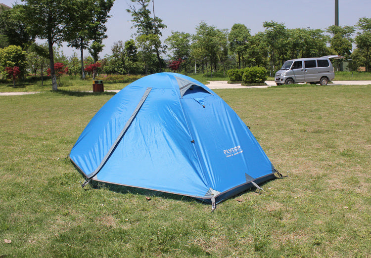 An eprolo Outdoor Double Camping Rainproof Tent is pitched on a grassy field beneath a clear sky, encircled by trees, with a gray van in the background.