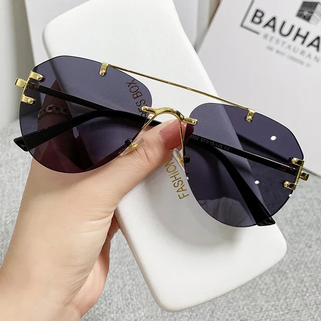 A hand holds stylish Vintage Rimless Alloy Gradient Pilot Sunglasses by eprolo, featuring an oversized black and gold frame with gradient lenses. They rest on a white rectangular case labeled FASHION, combining style with eye protection. The blurred background shows text and a box.