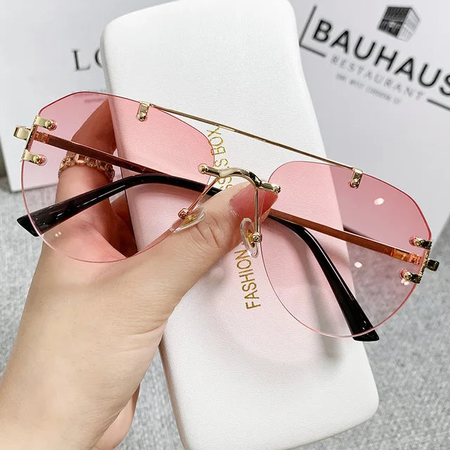 A hand holds eprolos Vintage Rimless Alloy Gradient Pilot Sunglasses, featuring pink-tinted lenses and gold rims. They rest on a white case with FASHION B partially visible. In the background, blurred text like BAUHAUS and RESTAURANT adds an artistic touch.