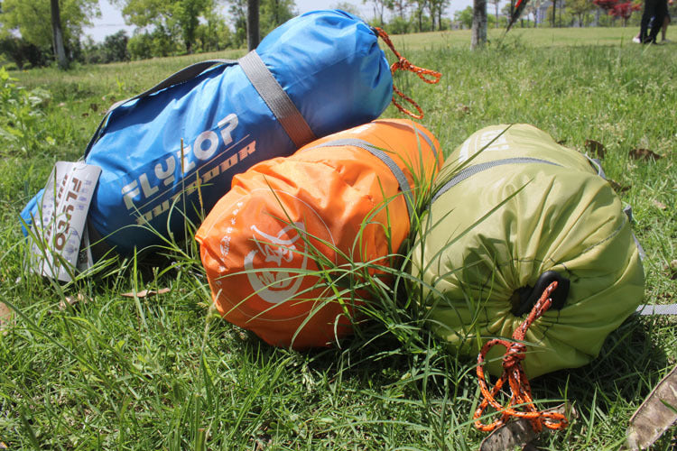 Three rolled-up sleeping bags in blue, orange, and green are on the grass beside an eprolo Outdoor Double Camping Rainproof Tent. The bags are strapped and partly covered by grass with blurred trees and greenery in the background.