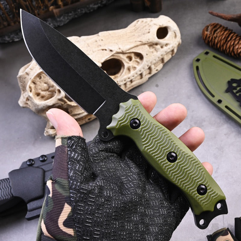 A hand in a camouflage glove holds the eprolo A3 High Hardness Outdoor Multifunctional Portable Knife, featuring a textured green handle and black blade. The stone-like object and sheath on the gray surface highlight its versatility as a survival tool.