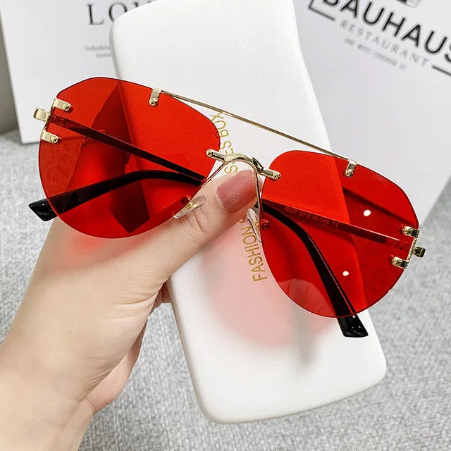 A hand holds Vintage Rimless Alloy Gradient Pilot Sunglasses from eprolo, featuring bold red lenses and gold frames, over a white Fashion Glass Box case. The background hints at a branded setting with indistinct text.