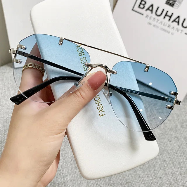 A hand holds eprolos Vintage Rimless Alloy Gradient Pilot Sunglasses with blue-tinted lenses and black temples, offering stylish eye protection. A white case and a Bauhaus Restaurant sign are seen in the background.