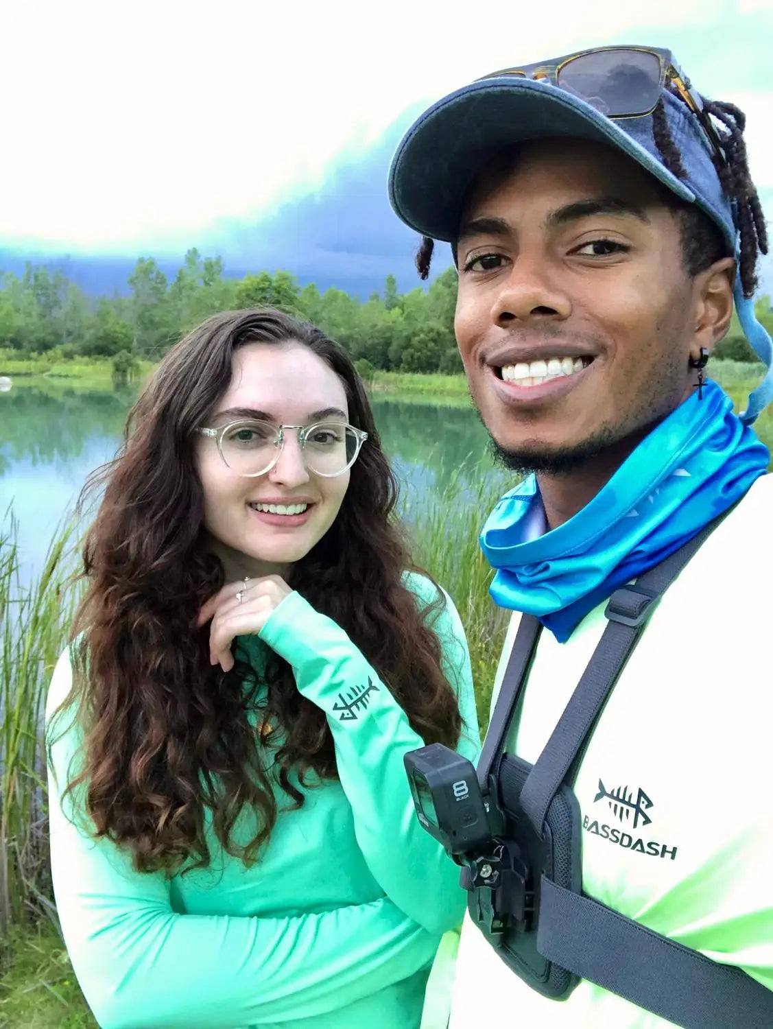 A smiling man and woman enjoy the outdoors near a pond. The man sports a cap, sunglasses, a bright moisture-wicking shirt, and a camera harness. The woman wears glasses, has long curly hair, and dons the vibrant Ten Essentials Bassdash Womens UPF 50+ Long Sleeve Performance Shirt. Its cloudy.