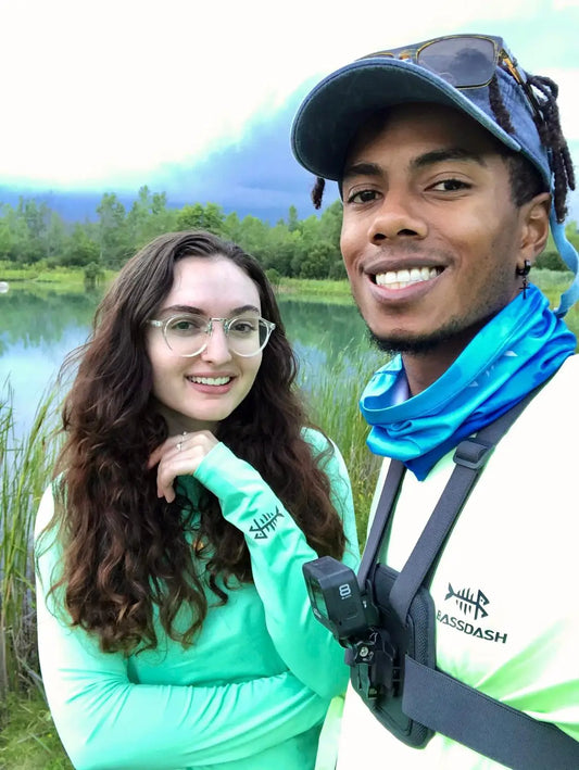 A smiling man and woman enjoy the outdoors near a pond. The man sports a cap, sunglasses, a bright moisture-wicking shirt, and a camera harness. The woman wears glasses, has long curly hair, and dons the vibrant Ten Essentials Bassdash Womens UPF 50+ Long Sleeve Performance Shirt. Its cloudy.