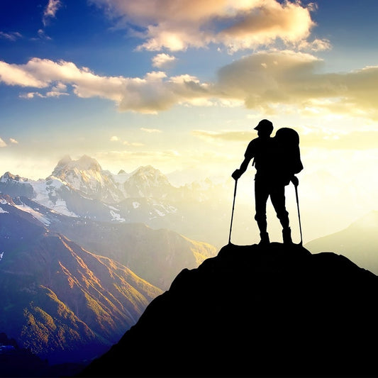 A hiker with Modalysts Adjustable Aluminum Trekking Poles stands on a mountain peak against majestic mountains and a vibrant sky at sunrise or sunset, capturing the essence of thrilling outdoor adventures.