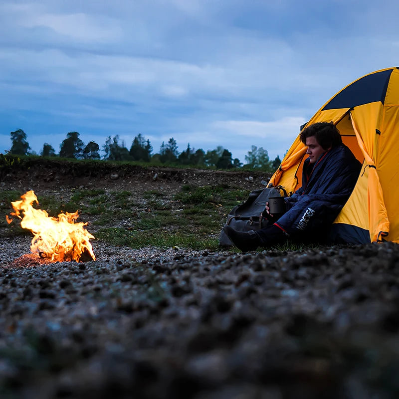 A person sits at the entrance of a yellow tent, wrapped in a Ten Essentials Widesea Ultralight Down Sleeping Bag. Surrounded by a cloudy sky and cool evening air with rustling trees nearby, they gaze at the campfire, staying snug and dry thanks to the waterproof fabric.