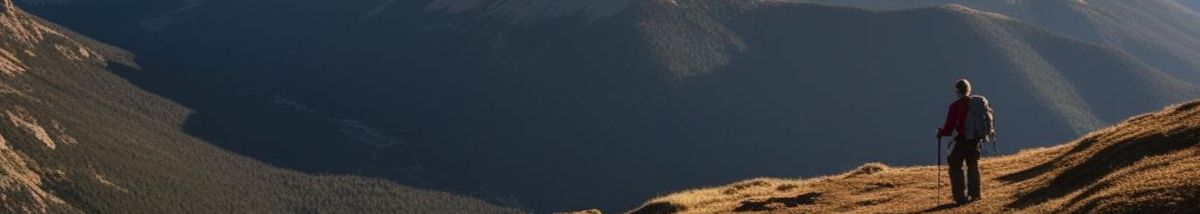 A hiker in a red jacket stands on a grassy ridge, overlooking a vast mountain landscape with deep shadows and valleys. The hiker looks towards the expansive view, suggesting a peaceful moment in nature.