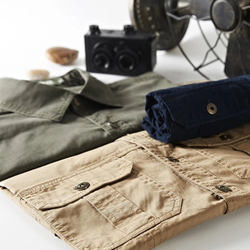 Two neatly folded Men’s Quick-Dry Tactical Shirts, one beige and one olive-green by Ten Essentials, sit on a surface. In the background are a black camera, rolled-up navy fabric, and a vintage fan adding visual interest.