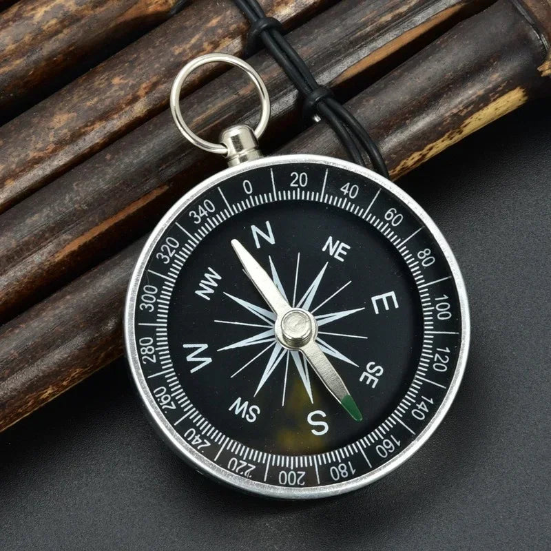 The Ten Essentials Portable Aluminum Emergency Compass, a silver-framed survival tool with a black dial featuring cardinal directions and degree markings, includes a subtly glowing fluorescent pointer and a metal loop. It rests beside wooden sticks on a dark surface.