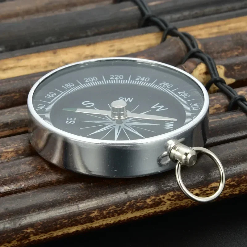 A Ten Essentials Portable Aluminum Emergency Compass sits on wood, showcasing cardinal directions and degree markings. Its silver tone, black face, fluorescent pointer, and side loop make it a perfect lightweight survival navigation tool for outdoor adventures.