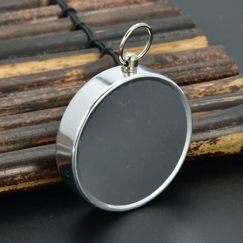 The Ten Essentials Portable Aluminum Emergency Compass, a round silver pendant, rests on parallel wooden sticks against a dark backdrop. Its reflective surface and small metal loop allow easy threading on a chain.
