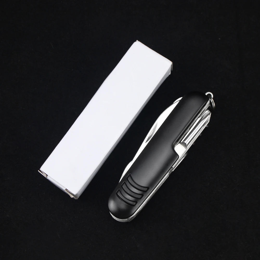 The Ten Essentials 11-in-1 Swiss Folding Pocket Knife with a black handle sits next to its white rectangular box on a black background, highlighting its stainless steel multitool features.
