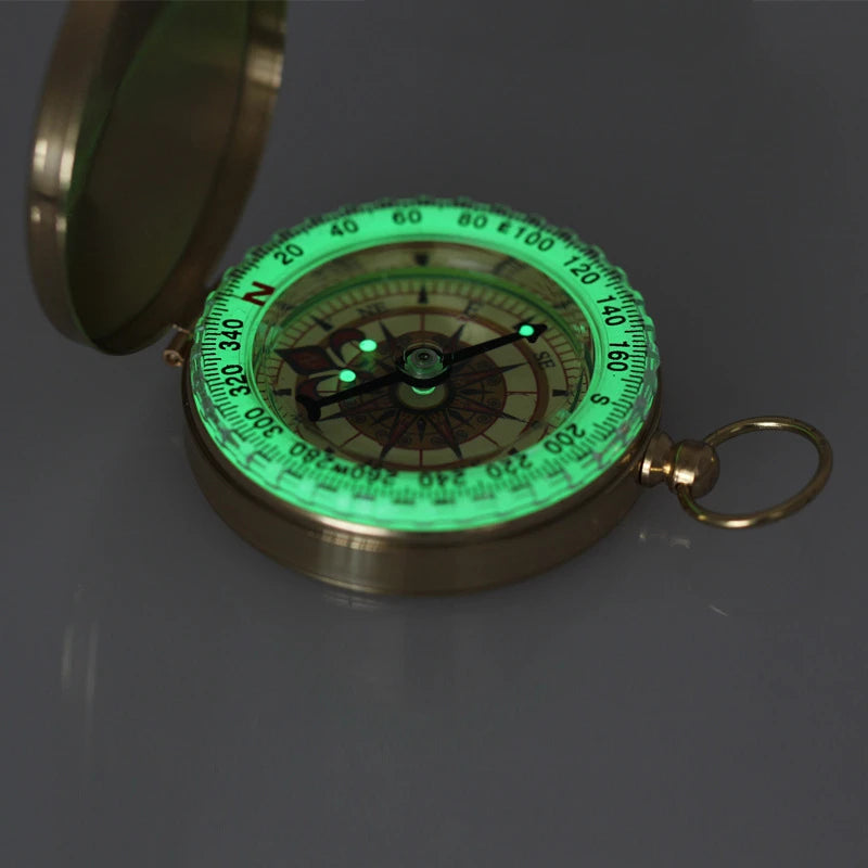 The Ten Essentials Portable Water-Resistant Luminous Brass Compass is perfect for outdoor adventures. It features a glow-in-the-dark green dial on a gray surface, displaying cardinal directions, degrees, and a decorative design. Ideal for survival situations, it comes with an attached small ring.