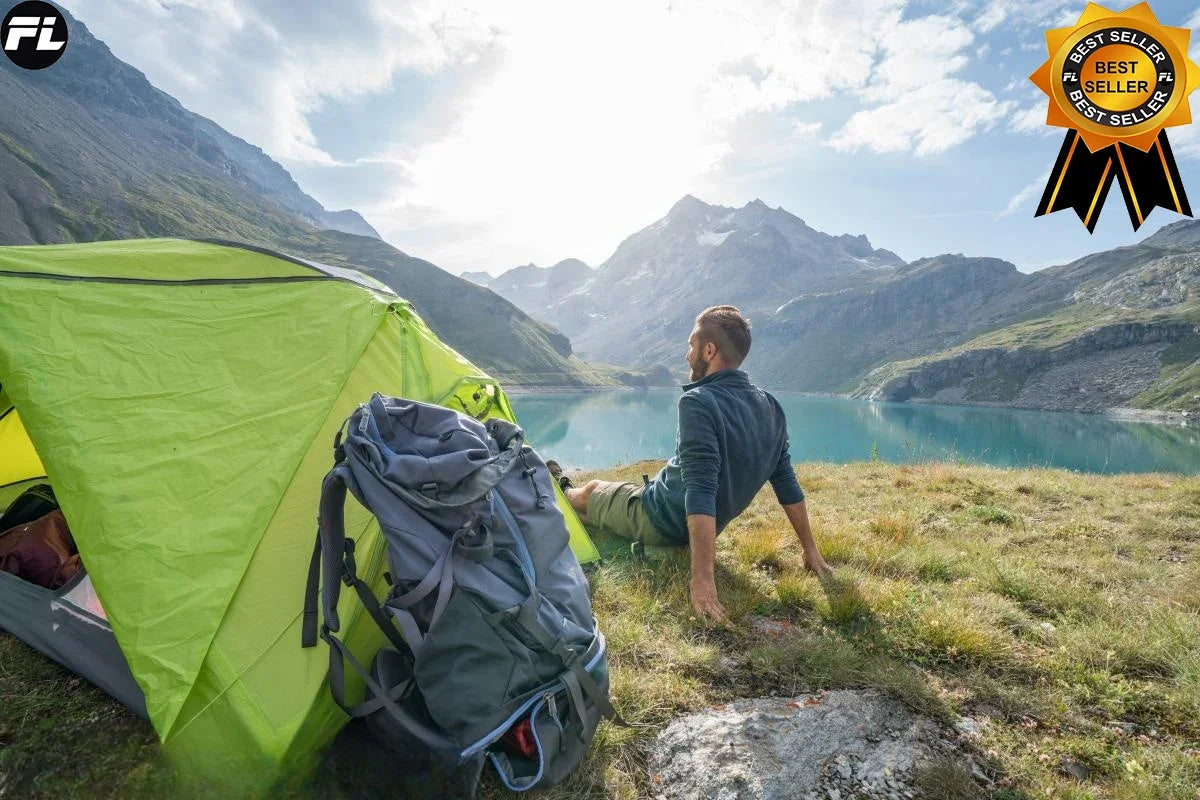 A man sits by a green camping tent, overlooking a serene mountain lake surrounded by high peaks under partly cloudy skies. Beside his backpack rests the Ten Essentials Folding Stainless Steel Pocket Knife, a portable survival and outdoor tool. A Best Seller badge is in the top right corner.