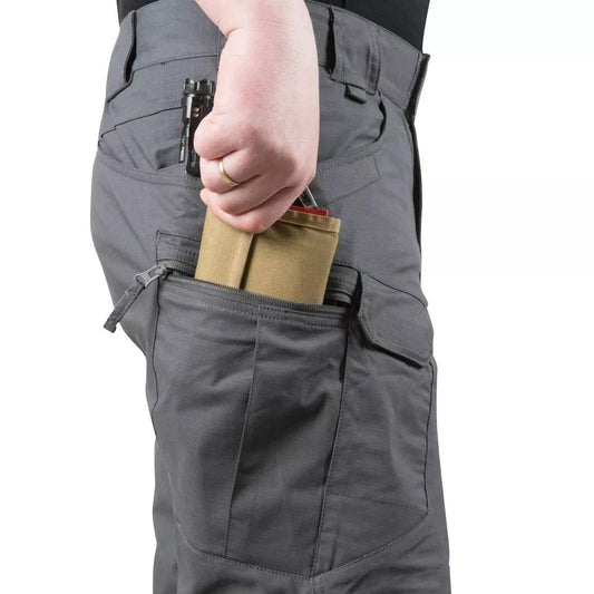 Dressed in Ten Essentials Men’s Tactical Cargo Shorts – waterproof and made from breathable cotton canvas, a person slips a tan wallet into a zipped side pocket. The multi-pocket design holds a black object clipped to the gray fabric, while they confidently wear a black top.
