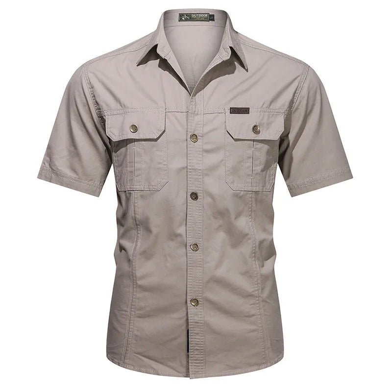The Men’s Quick-Dry Tactical Shirt by Ten Essentials is a beige, short-sleeve button-up crafted from breathable cotton, featuring two front pockets with button closures. Its collar and stitching details offer a military-inspired, structured, yet casual look.
