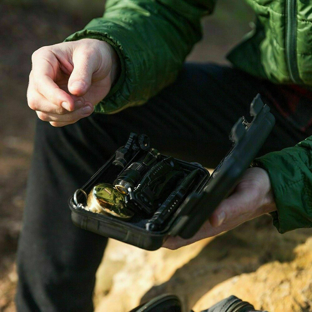 Seated outdoors under the sunlight, a person in a green jacket holds a small Modalyst 14-In-1 Outdoor Emergency Survival Kit packed with tactical gear, including various tools and a compass. Shadows are cast on the ground as they stay prepared.
