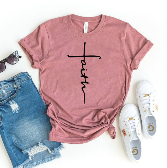 Flat lay of T-Shirt Crazeds pink Faith T-shirt featuring a black cursive cross design, offering premium fabric and unisex fit. Paired with distressed denim shorts, white sneakers, and sunglasses on a crisp white background.