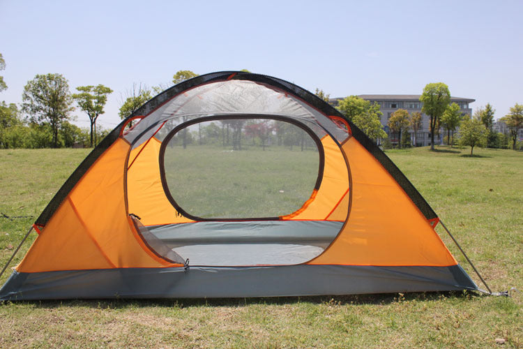 An eprolo Outdoor Double Camping Rainproof Tent in orange and gray is set up on a grassy field, ideal for outdoor camping. Trees and a building complement the clear blue sky in the background.