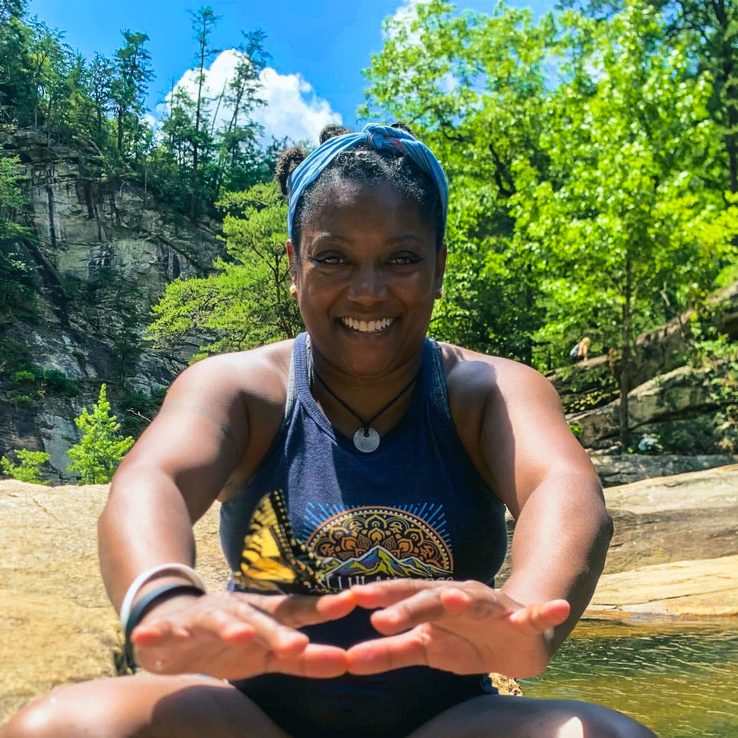 A person with a joyful expression sits outdoors on a rock near a stream, surrounded by lush greenery and a cliff. They wear a blue sleeveless top and a headband, with a butterfly visible on their hand. The background shows a bright blue sky.
