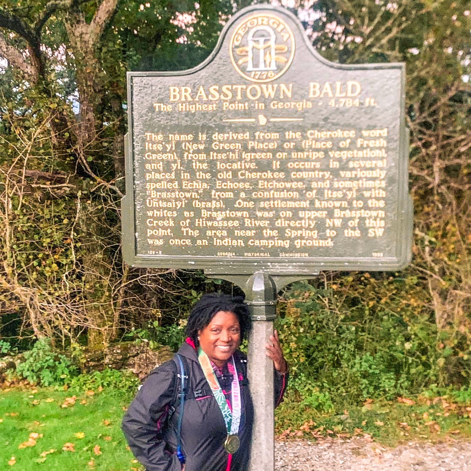 A person stands smiling in front of a historical marker titled Brasstown Bald, the highest point in Georgia. The sign explains the names Cherokee origins. Trees and greenery surround the area.