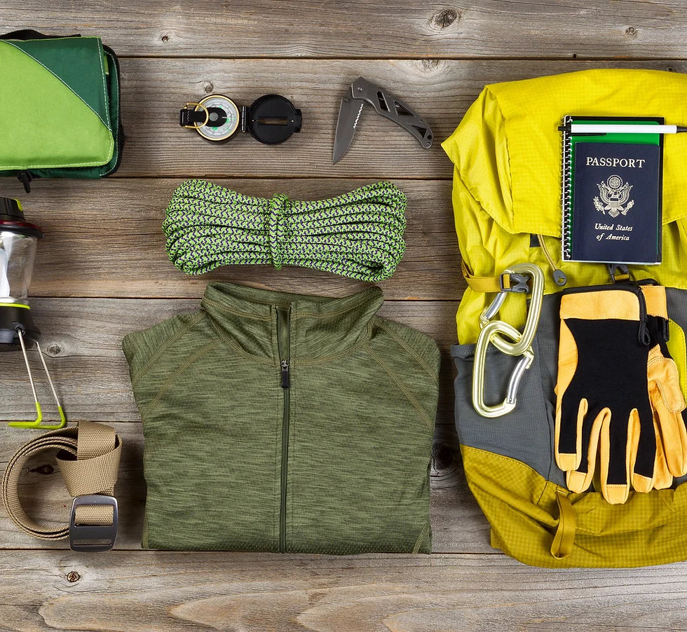A flat lay of travel and outdoor gear on a wooden surface. Includes a yellow backpack, passport, compass, knife, rope, green jacket, gloves, headlamp, flashlight, and water bottle. Items are neatly organized with a green and yellow color theme.
