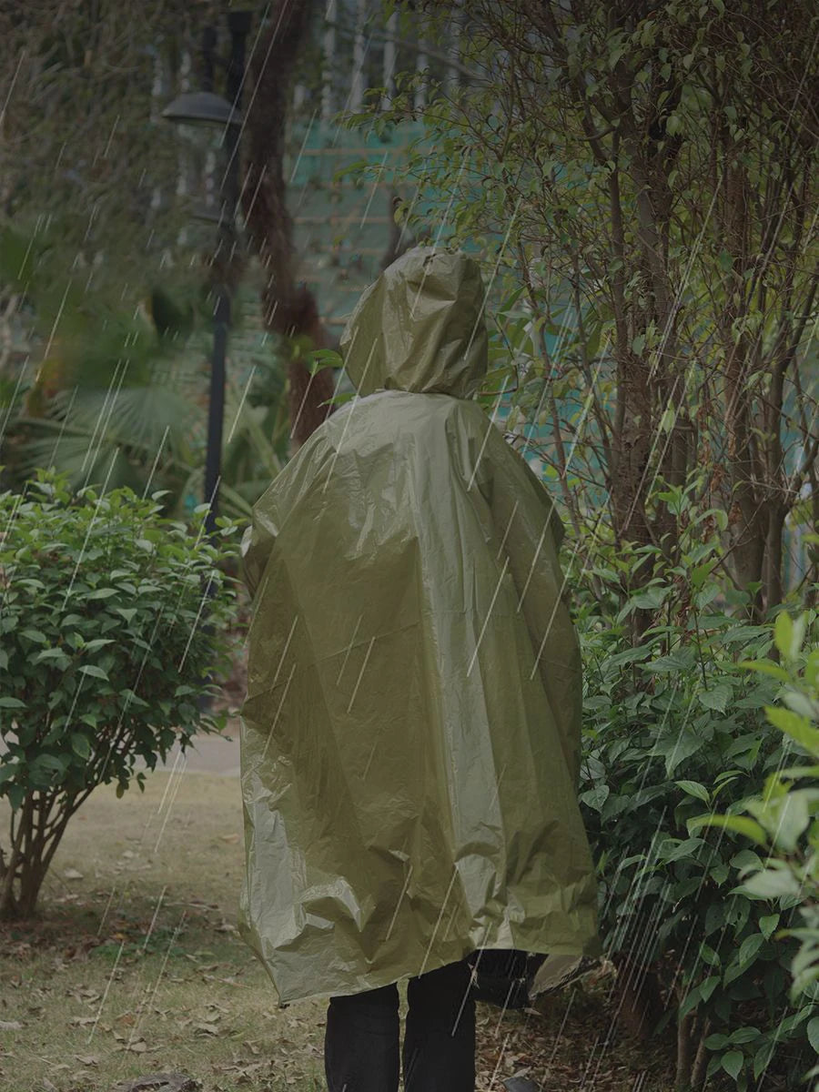 A person clad in a Ten Essentials Emergency Rain Poncho with Mylar Blanket Liner stands in the rain, surrounded by lush bushes and trees outdoors, as droplets visibly fall.