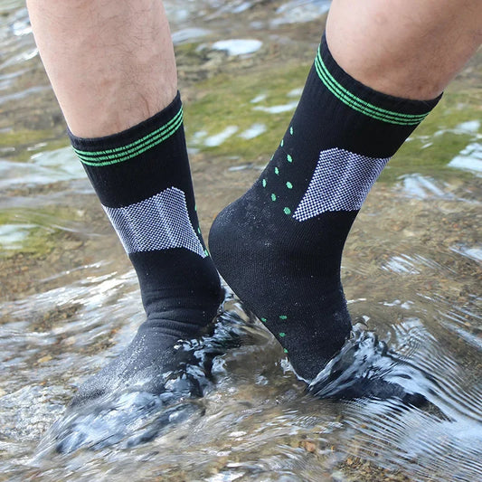 A person wearing Ten Essentials Waterproof Outdoor Sports Socks, featuring green and gray patterns, stands in shallow water with feet submerged but dry. The breathable socks resist water, perfect for hiking and skiing, while the rocky streambed shows beneath.