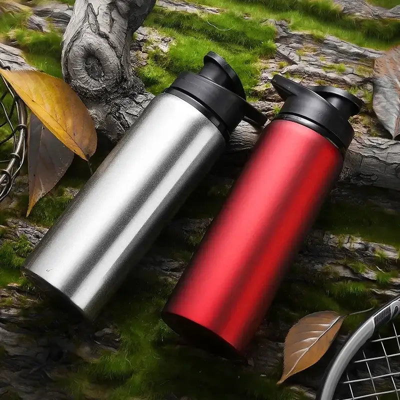 Two Ten Essentials Portable Stainless Steel Water Bottles – 700ml Sport & Travel Flasks, in silver and red, rest on a mossy log. Fallen leaves and branches are scattered about, creating an ideal setting for outdoor sports enthusiasts.