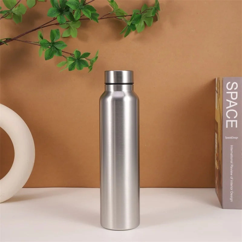 The 1000ml Stainless Steel Sports Water Bottle by Ten Essentials, known for its durability and eco-friendliness, stands on a white surface with a looping ceramic vase on the left and a book titled SPACE on the right. Green leaves hang above against a brown background, enhancing its natural appeal.