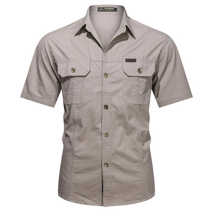 The Men’s Quick-Dry Tactical Shirt by Ten Essentials is a beige, short-sleeve button-up crafted from breathable cotton, featuring two front pockets with button closures. Its collar and stitching details offer a military-inspired, structured, yet casual look.